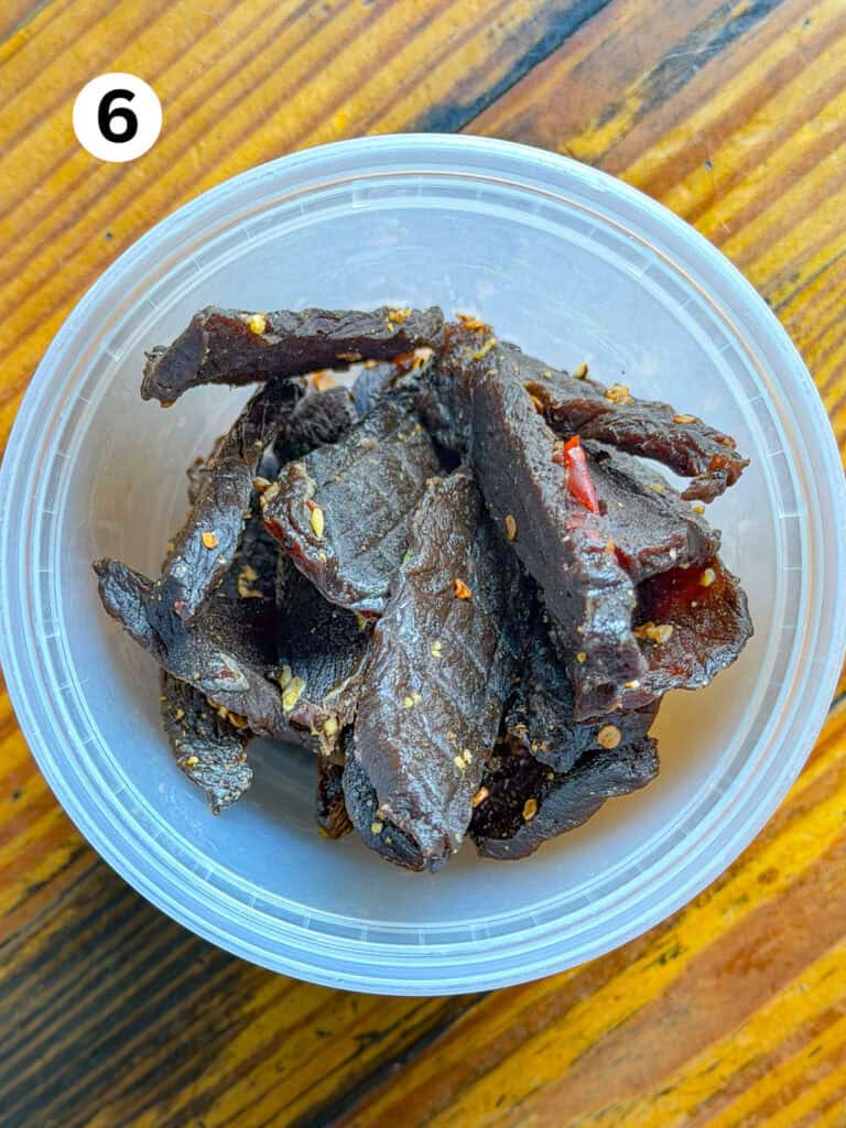 Venison jerky in a plasic storage container on a wooden table.