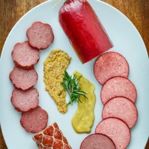 Venison summer sausage on a white plate with mustard and rosemary.