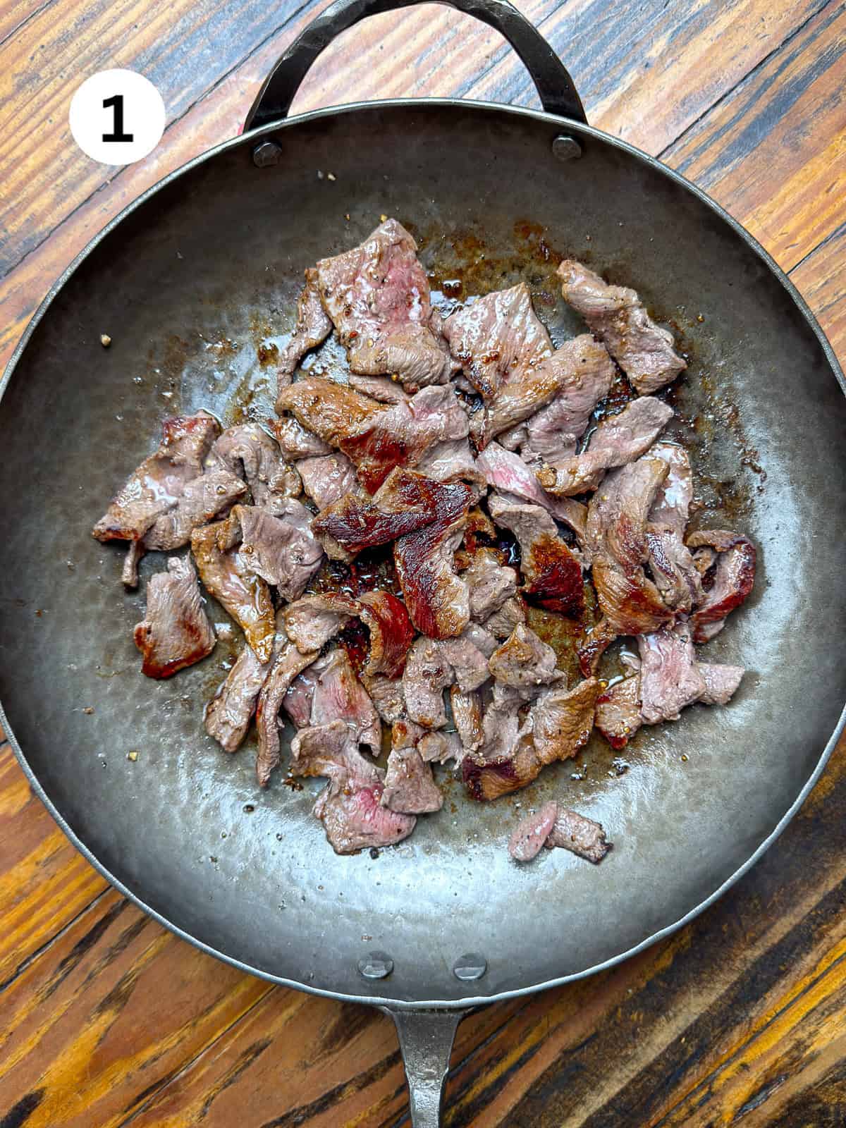 Seared sliced venison in a carbon steel pan on a wooden table.